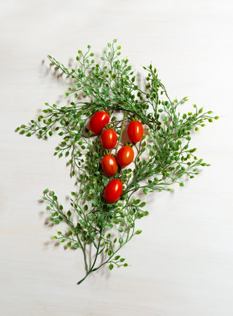 Still life of elongated branch of red tomatoes among small green leaves on white wooden table. Vertical.の写真素材