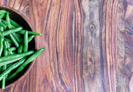 Wooden bowl of fresh green beans on left side of wooden cutting board, with copy space, lit with natural sunlightの写真素材