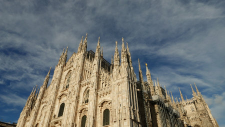 The towering spiers of Milan Cathedral reach towards the sky, surrounded by wispy clouds, showcasing intricate details against the bright backdrop of morning light.の写真素材