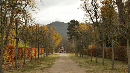 A tranquil park pathway surrounded by trees in autumn colors leads toward a decorative fountain. The nearby mountains add a scenic backdrop to this peaceful setting.の写真素材