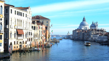 Boats glide along the Grand Canal, surrounded by stunning historic buildings. The blue sky casts a serene light over the water, depicting a tranquil day in Venice.の写真素材