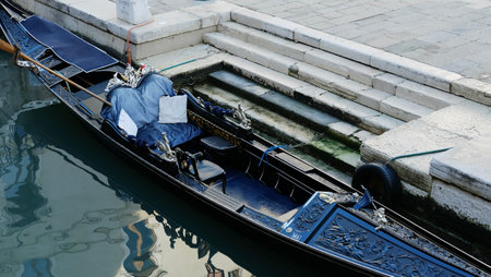 A gondola with intricate designs is tied to the shore, partially covered, and surrounded by calm water in Venice. The peaceful setting suggests early morning tranquility in the city.の写真素材