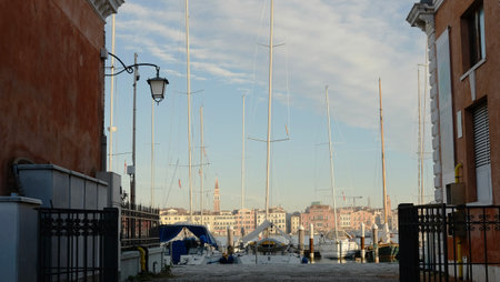A picturesque waterfront view captures several sailboats moored in a marina, framed by charming buildings. The scene is alive with the warm glow of afternoon light and clear skies.の写真素材