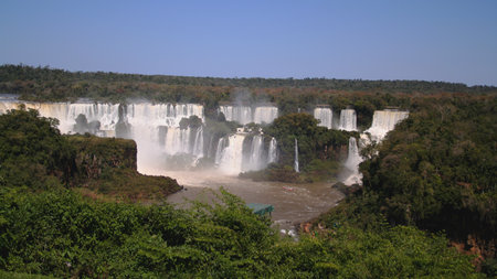 Iguazu Falls showcases its breathtaking beauty with numerous waterfalls flowing into the river. Lush greenery surrounds this natural wonder, highlighting its majesty under a clear blue sky.の写真素材