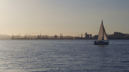 A sailboat navigates calmly across the water during sunset, with silhouettes of cranes and buildings creating a picturesque backdrop against the fading light.の写真素材