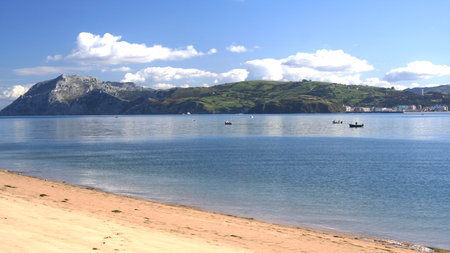 A serene beach features soft sand and clear blue waters. Small boats are scattered on the calm water, with hills and a town in the background under a sunny sky.の写真素材
