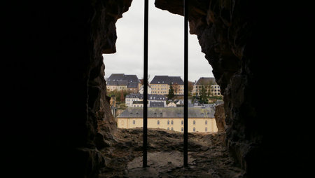 Peering through a stone opening in the fortress, a mix of old and new architecture is visible. The cloudy sky adds a touch of mystery to the cityscape below.の写真素材