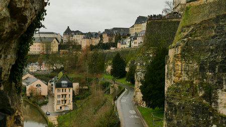 Luxembourg presents a stunning view of rocky cliffs and charming buildings. A winding road passes through the valley, with visitors on the cliff's edge taking in the scenery on a cloudy day.の写真素材