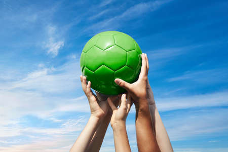 Group of football fans holding red soccer ball in green cloudy sky. Kid s and adult s hands together hold ballの写真素材