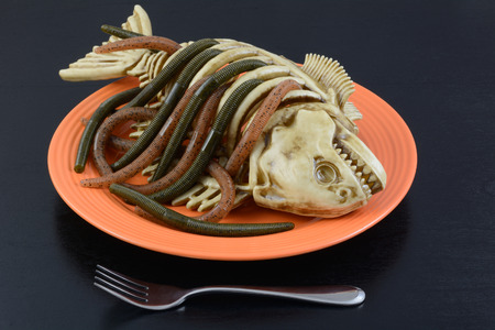 Halloween dinner dish worms crawling out of fish skeleton on orange plate with fork on black tableの写真素材