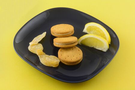 Cookies with lemon ginger cream filling  with lemon wedges and crystallized ginger on dark blue snack plate on yellow backgroundの写真素材