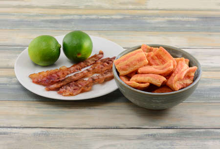 Puffed bacon chips flavored with zingy line in ceramic bowl with two fresh limes and strips on bacon...