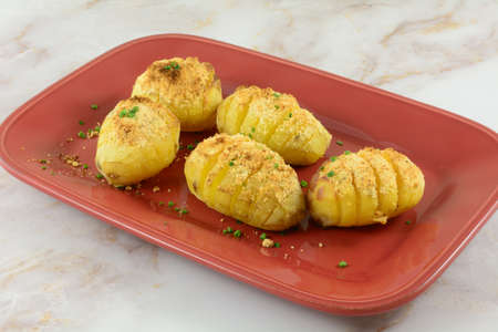Baked hasselback potatoes sprinkled with cheese on red serving plate