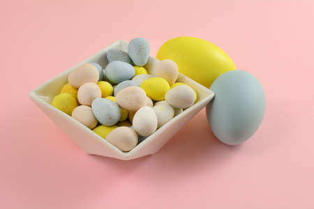 Pastel colored candy Easter eggs in white dessert bowl with two painted Easter eggs on pink...