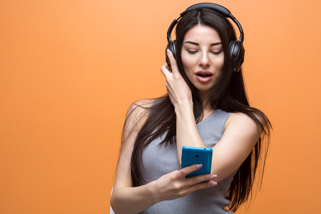 Portrait of a young beautiful girl listening to music through the headset and looking at her smart phone against orange background. Lifestyle, people and technology conceptの写真素材