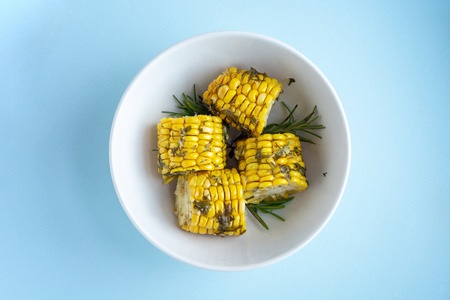 Cooked in the oven corn with herbs in white bowl closeup over blue background, top view, copy space...