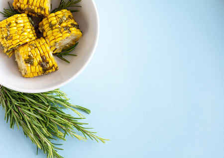Cooked in the oven corn with herbs in white bowl and rosemary flat lay over blue background, top view, copy space for text.の写真素材