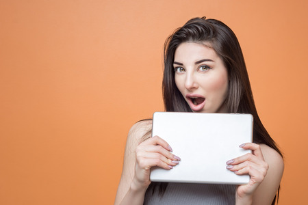 Portrait of a young excited brunette girl with tablet in her hands looking at the camera with open mouth against orange background. Lifestyle, people and technology concept.の写真素材