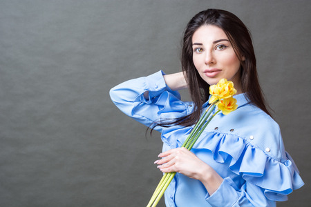 Portrait of a young beautiful brunette woman in blue shirt holding yellow flowers in her hands and looking at the camera against grey background.の写真素材