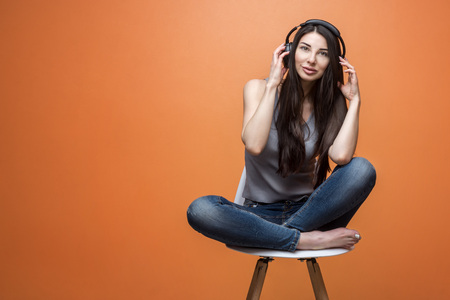 Beautiful casual brunette in denim listening to music in headphones and holding her hands near her head, sitting in a chair on orange backgroundの写真素材
