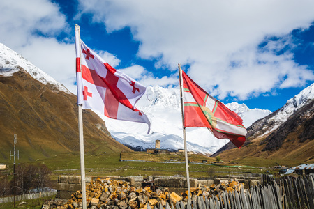 Picturesque landscape of LaMaria church in aged rural community Ushguli against beautiful mountains with snowy peaks, Mt. Shkhara, the highest peak of Georgia on the background, Georgian and Svaneti flags in the foregroundの写真素材