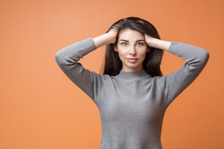Closeup portrait of a young beautiful brunette woman with long hair holding her hands in her hair and looking to the camera, thinking about something against orange background.の写真素材