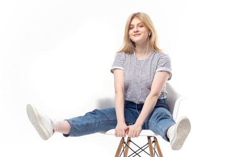 Pretty blond woman in denim and sneakers sitting on chair with legs apart while listening to music on white backgroundの写真素材