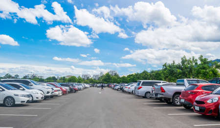 Car parking in large asphalt parking lot with trees, white cloud and blue sky background. Outdoor parking lot with fresh ozone and green environment of transportation and modern technology conceptの写真素材