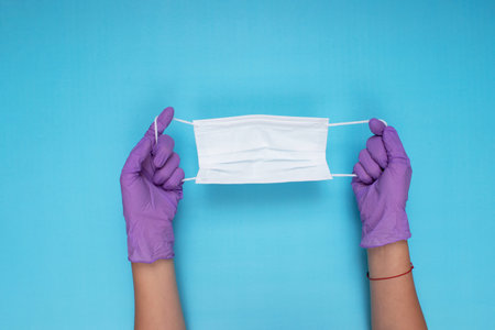 doctor wearing a white surgical mask and a pair of lilac colored gloves on a light blue background