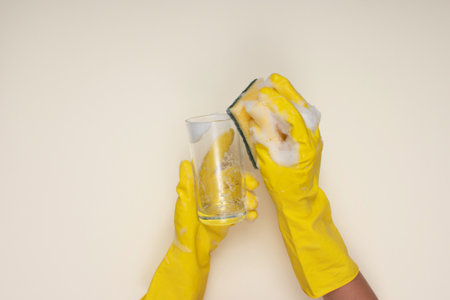 woman wearing yellow cleaning gloves on light color background cleaning glass cup with cleaning...