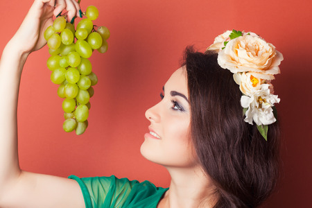 beautiful young woman wearing wreath and holding green grapes against red backgroundの写真素材