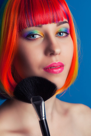 beautiful woman wearing colorful wig and holding make-up brush against blue backgroundの写真素材