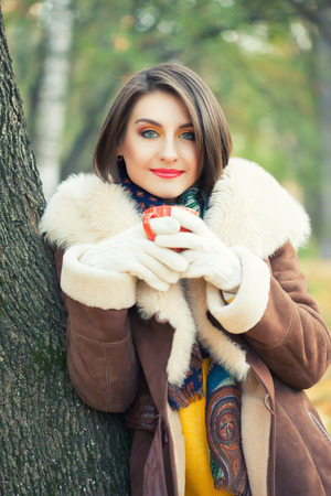 Beautiful young woman in autumn park with cup in handsの写真素材