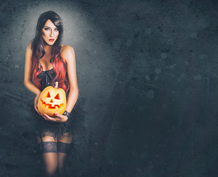Beautiful witch in black gothic Halloween costume holding Jack-o-lantern in hands on grunge backgroundの写真素材