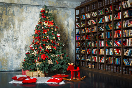 a cozy, festive scene featuring a beautifully decorated Christmas tree placed beside a large bookshelf. The tree is adorned with an abundance of ornaments, mostly in red, gold, and white, with decorative flowers, baubles, ribbons, and lights, creating a vibrant holiday atmosphere. Beneath the tree, there are wrapped presents and festive red pillows with white designs, placed on a soft white rugの写真素材