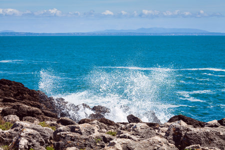 A peaceful ocean scene with shimmering blue water stretching to the horizon, rocky formations in the foreground, and a lone rock emerging from the waves, creating a tranquil and serene coastal atmosphereの写真素材