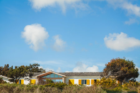 A bright, cozy house with yellow shutters nestled among trees and greenery under a vast blue sky with scattered clouds, creating a peaceful and inviting countryside settingの写真素材