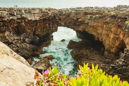 A dramatic natural arch carved into the rocky cliffs, with turquoise waves crashing beneath, surrounded by rugged terrain and vibrant coastal plants, creating a striking contrast of textures and colorsの写真素材