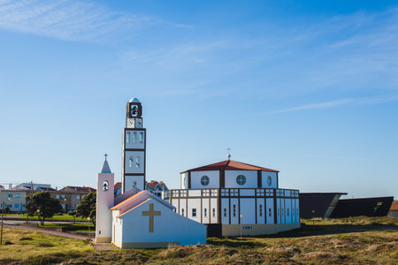 a modern church building located in a peaceful, rural area with clear blue skies above. The church has a unique architectural design, with clean, white walls accented by brown lines framing the structure. A tall clock tower, with an additional bell tower, stands prominently next to the main circular building.の写真素材