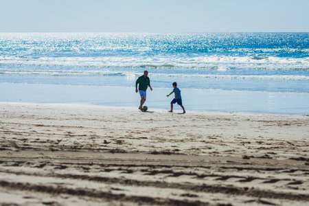 In this image, a man and a young boy are playing soccer on a sandy beach. The man, wearing a green shirt and shorts, is gently guiding the ball as the boy, dressed in blue, reaches out, possibly trying to intercept or pass the ball. The sun reflects brightly off the ocean in the background, with waves rolling towards the shore.の写真素材
