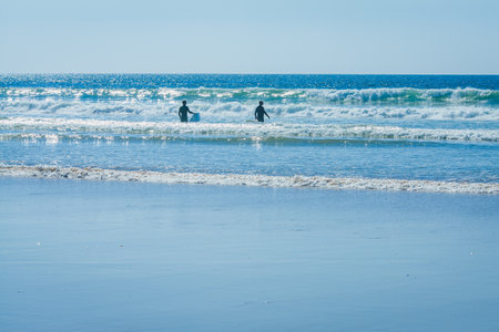 This image shows two surfers wading into the ocean, walking through the shallow water as they head...