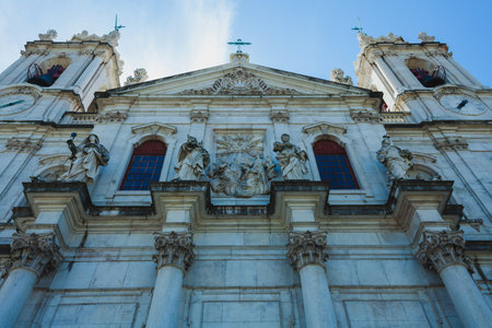 the grand faÃ§ade of a historic church or cathedral, featuring intricate architectural elements and several sculptures adorning the structure. The stone building is characterized by tall columns with Corinthian capitals and an array of statues placed in niches or on ledges. Above, a detailed relief in the center depicts a religious scene, possibly of heavenly figures. The building's towering spires and arched windows, framed by ornate carvings, contribute to the imposing and majestic appearance of the structure.の写真素材
