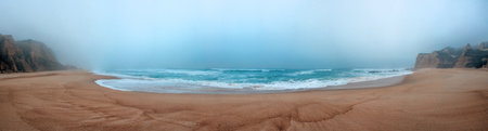 A serene, foggy beach scene with gentle waves rolling onto the shore and towering cliffs in the background, creating a tranquil and atmospheric landscapeの写真素材