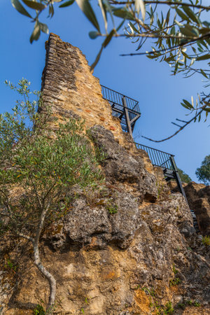 A stone wall ruin with a metal railing and staircase clings to a rocky cliff face, framed by olive tree branches against a clear blue skyの写真素材