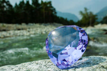 A brilliant faceted purple gemstone rests on a rock with a scenic river and forested mountains in...