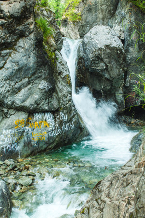 This image captures a small but vibrant waterfall cascading between rugged rocks into a pool of clear, green-blue water. The rocks around the waterfall are steep and jagged, adding to the wild, untamed feeling of the sceneの写真素材