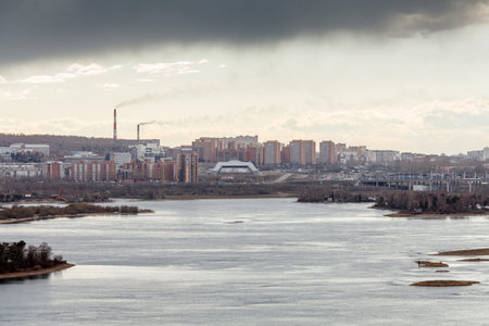 A cityscape view across a wide river, featuring tall residential buildings and industrial smokestacks under a cloudy skyの写真素材
