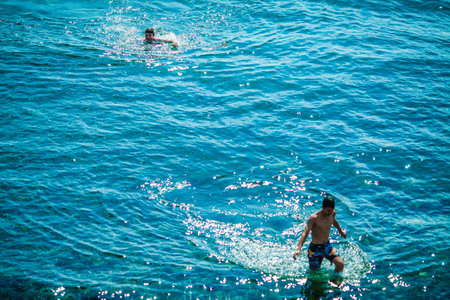 An overhead view of two people swimming and splashing in the vibrant blue water, one fully immersed while the other wades through the shallows, with sunlight sparkling off the water's surfaceの写真素材