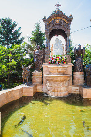 A beautifully ornate fountain with sculptures surrounding a religious monument, reflecting in a serene pond with swimming fish in a sunny dayの写真素材