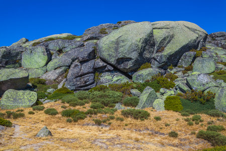 A rugged mountain landscape with large boulders, sparse vegetation, and a distant dam under a clear blue skyの写真素材
